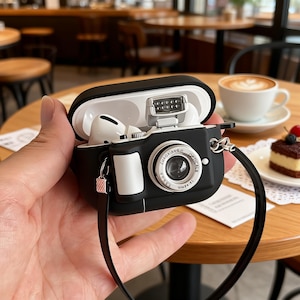 May include: A black and white camera-shaped case for wireless earbuds, with a black strap. The case is open, showing white earbuds. The case has a lens and flash details, with text on the lens.