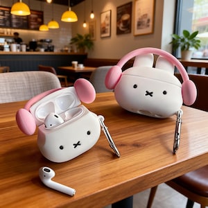 Op de afbeelding: Twee witte en roze AirPod-hoesjes met konijnenthema en een enkele AirPod liggen op een houten tafel. De hoesjes hebben zwarte ogen en een zwart kruis als mond. De hoesjes hebben roze koptelefoons. Eén hoesje is open en onthult de witte AirPods binnenin.