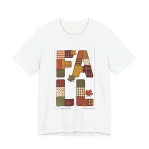 May include: A white short-sleeve t-shirt featuring the word FALL printed in a patchwork design. Each letter is composed of various fabric patterns in shades of orange, brown, green, and yellow, accented with small brown and orange maple leaf illustrations.