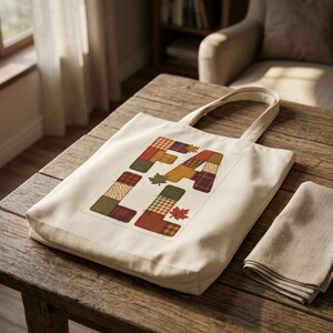 May include: A cream-colored canvas tote bag featuring the word FALL printed in large, block letters. Each letter is composed of various patchwork-style patterns in shades of orange, brown, green, and red, accented by small maple leaf illustrations.