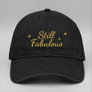 Puede incluir: Gorra de béisbol negra con texto bordado dorado "Still Fabulous". La gorra tiene una visera curva y un diseño de seis paneles. El texto está acompañado de tres pequeñas estrellas.