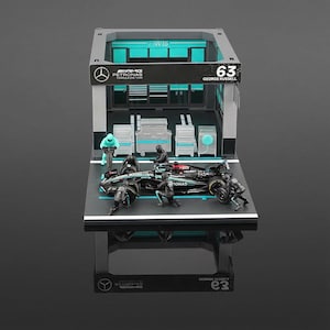 Puede incluir: Una maqueta de garaje de F&oacute;rmula 1 con un coche de carreras negro y turquesa con el n&uacute;mero 63. La escena incluye figuras en miniatura, cajas de herramientas y el logotipo de AMG Petronas. El garaje tiene una combinaci&oacute;n de colores gris y turquesa.
