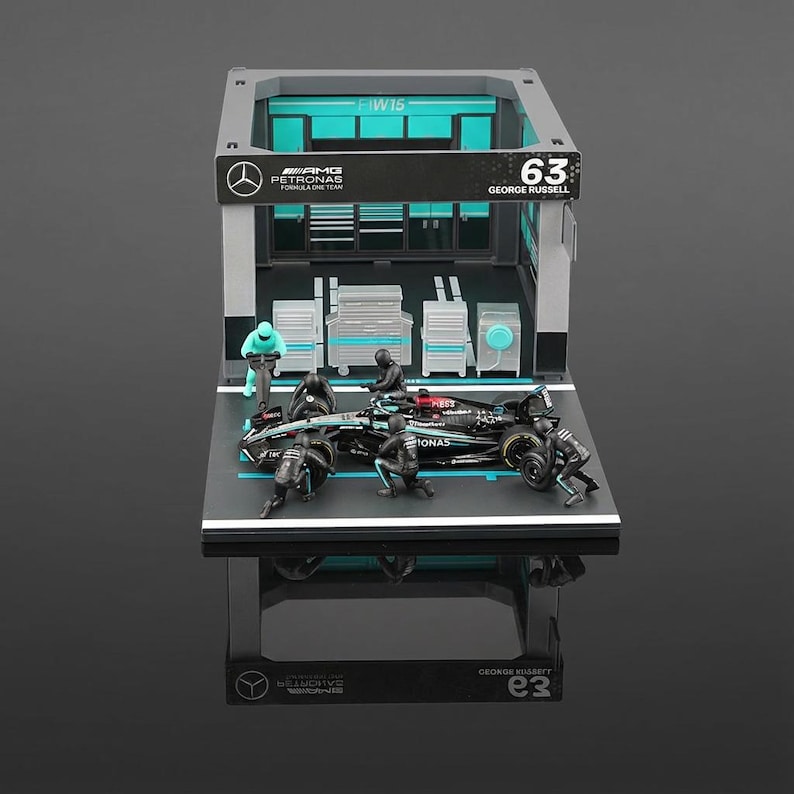 Puede incluir: Una maqueta de garaje de F&oacute;rmula 1 con un coche de carreras negro y turquesa con el n&uacute;mero 63. La escena incluye figuras en miniatura, cajas de herramientas y el logotipo de AMG Petronas. El garaje tiene una combinaci&oacute;n de colores gris y turquesa.