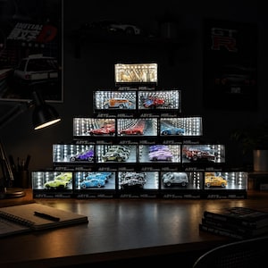 Puede incluir: Una exhibición de modelos de coches en miniatura en vitrinas iluminadas. Las vitrinas están dispuestas en forma de pirámide, con coches de varios colores y estilos visibles. El texto "ABYSAL MIRROR" está impreso en las vitrinas.