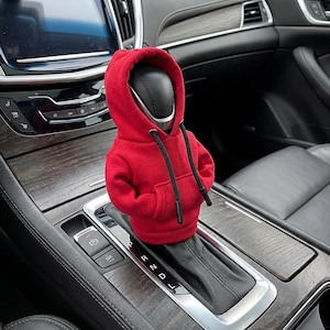 May include: A red miniature hoodie covers a car's gear shift. The hoodie has a drawstring and a kangaroo pocket. The gear shift is black and silver, and the car's interior features wood grain and black leather.