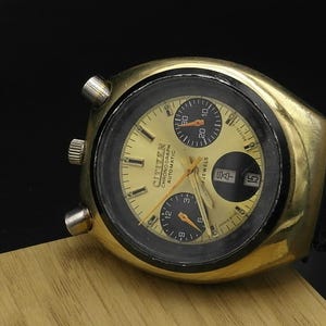 Op de afbeelding: Een vintage Citizen chronograaf automatisch horloge in goudkleur met een zwarte lunette en een gouden wijzerplaat. Het horloge heeft drie sub-wijzerplaten en een datumvenster. Het horloge heeft een zwarte leren band.