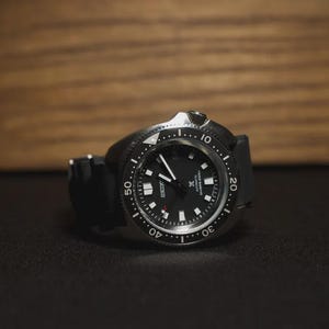May include: A black wristwatch with a black band and a silver bezel. The watch face is black with white markers and hands. The watch is set against a dark background with a wooden backdrop.