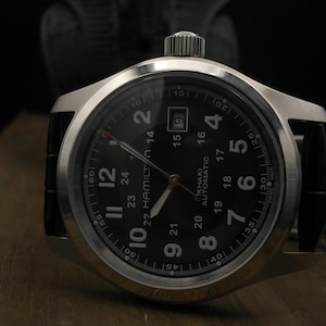 May include: Close-up of a silver-toned wristwatch with a black face and a black leather band. The watch face displays the time with white numerals and hands. The watch has the text "HAMILTON KHAKI AUTOMATIC".
