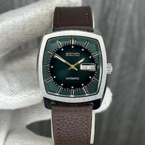 Könnte beinhalten: Eine Vintage Seiko Automatik-Uhr mit dunkelgrünem Zifferblatt, goldenen Akzenten und einem braunen Lederarmband. Das Zifferblatt zeigt Tag und Datum an. Die Uhr hat ein silberfarbenes Gehäuse.