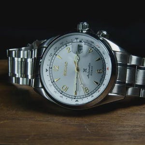 May include: A silver-toned Seiko watch with a light-colored face and a stainless steel band. The watch face features gold-colored numerals and hands, with the brand name and model details visible. The watch is displayed on a wooden surface.