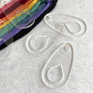 May include: Clear acrylic thread organizers with a teardrop design, alongside a rainbow of embroidery floss. The floss colors include red, orange, yellow, green, blue, purple, and black. The organizers have a small hole for hanging.