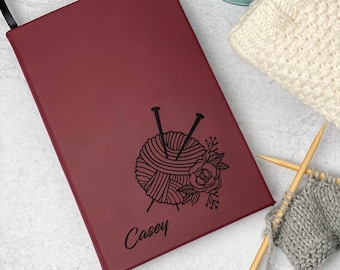Op de afbeelding: Een bordeauxrood notitieboek met een zwarte afbeelding van garen, naalden en bloemen, met de naam "Casey" eronder. Een zwart lint als bladwijzer is zichtbaar. Breinaalden en een kleine gebreide lap liggen op de achtergrond.