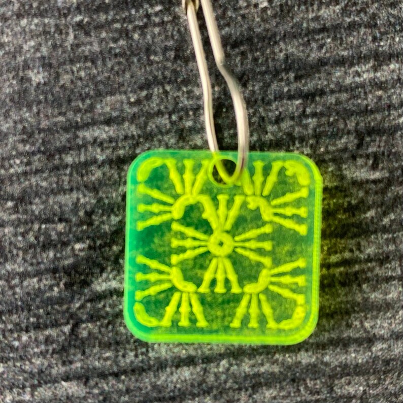 Pu&ograve; includere: Un ciondolo quadrato verde brillante con un designo decorativo, che presenta un motivo ripetitivo di linee radianti. Il ciondolo &egrave; attaccato a un anello di metallo argentato.