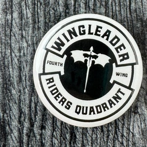 May include: White round sticker with black text that reads "Wing Leader Fourth Wing Riders Quadrant". A black and white illustration of a winged creature with a spear is in the center.