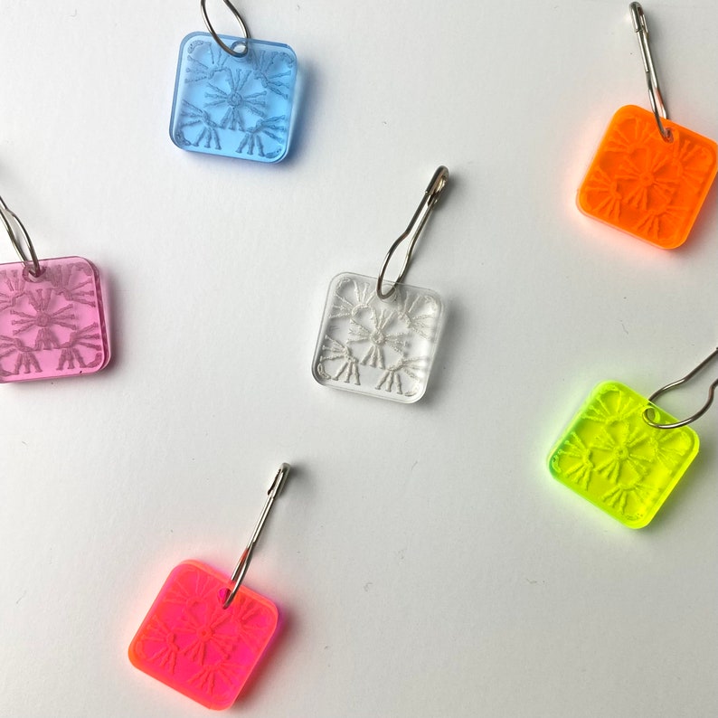 Pu&ograve; includere: Cinque marcapunti in plastica colorati con anelli metallici. I marcapunti sono quadrati e presentano un motivo a fiocco di neve. I colori sono rosa, blu, trasparente, arancione e giallo.