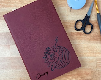 Op de afbeelding: Een bordeauxrood notitieboek met een bloemen- en garenbalontwerp, met de naam "Casey" gegraveerd op de kaft. Een lichtblauw meetlint, een zwarte schaar en een haaknaald liggen naast het notitieboek op een houten oppervlak.