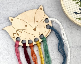 May include: A wooden fox-shaped thread organizer with seven holes for storing embroidery floss. The holes are numbered 1 through 7. The organizer is painted with a light brown stain and has a white background. The floss is in a variety of colors, including pink, red, orange, yellow, green, blue, and white.