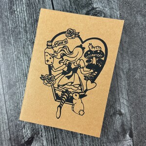 May include: A brown paper notebook featuring a black illustration of Alice in Wonderland holding a bottle labelled "Drink Me". The illustration is heart-shaped and includes a mushroom, a caterpillar, and a rabbit.