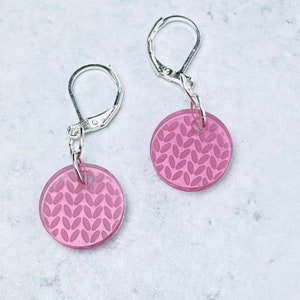 May include: A pair of pink earrings with a knit pattern. The earrings are made of acrylic and have silver hooks.