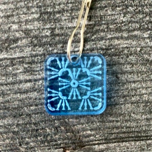 Pu&ograve; includere: Un ciondolo quadrato in plastica blu con un design decorativo. Il ciondolo &egrave; attaccato a un anello di metallo argentato.