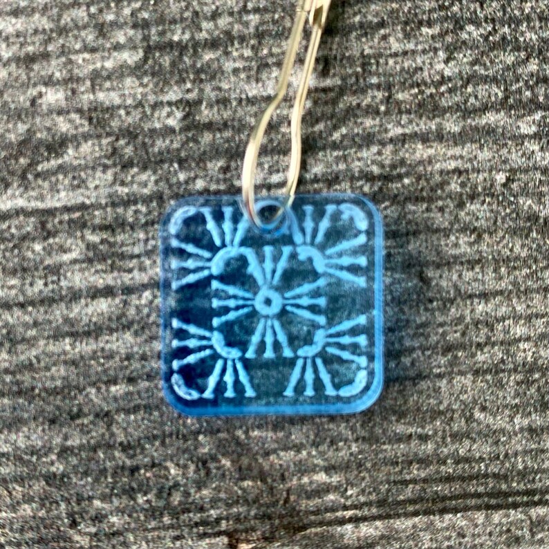 Pu&ograve; includere: Un ciondolo quadrato in plastica blu con un design decorativo. Il ciondolo &egrave; attaccato a un anello di metallo argentato.