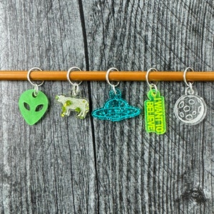 May include: Five colorful stitch markers with different designs, including a UFO, a cow, a heart, a moon, and a rectangle with the text "I want to believe".