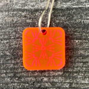 Pu&ograve; includere: Un ciondolo quadrato di colore arancione brillante con un design geometrico rosa. Il ciondolo &egrave; attaccato a un anello argentato.