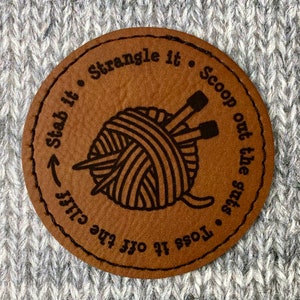 May include: A brown leather circle tag with black stitching and text that reads "Toss it off the cliff -> Stab it • Strangle it • Scoop out the guts". A black and white illustration of a ball of yarn with two knitting needles is in the center of the tag.