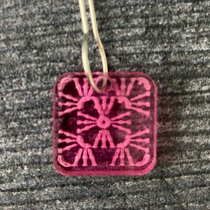 Pu&ograve; includere: Un ciondolo quadrato rosa con un designo dettagliato e intricato. Il ciondolo ha un foro nella parte superiore per essere attaccato a una collana o un braccialetto.
