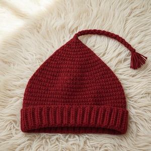 May include: A knitted burgundy hat with a pointed top and a braided cord ending in a tassel. The hat features a ribbed brim and is made of a soft, textured yarn. The hat is displayed on a fluffy, white surface.