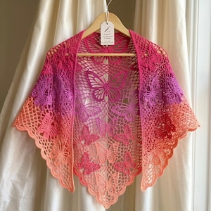 May include: A handmade, open-weave shawl in a butterfly design. The shawl features a gradient of colors, transitioning from peach to pink and purple. The shawl is displayed on a wooden hanger against a white backdrop.