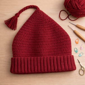 May include: A handmade, burgundy-colored crochet hat with a pointed top and a ribbed brim. The hat features a braided cord and tassel detail. Crochet hooks, yarn, stitch markers, and scissors are also visible.