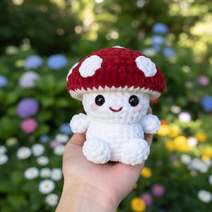 Op de afbeelding: Een gehaakte paddenstoel knuffel met een rode hoed en witte stippen. De paddenstoel heeft een lachend gezicht met zwarte ogen en roze wangen. De knuffel is wit en heeft kleine armen en benen.