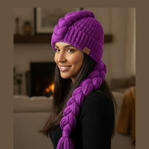 May include: A vibrant purple knit hat with a braided design, featuring a matching long braid that hangs down. The hat has a small brown tag on the side. The hat is a winter accessory.