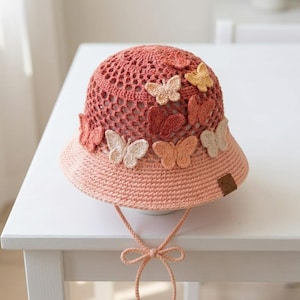 May include: A crocheted sun hat in shades of pink and coral, adorned with multiple butterfly appliqués in various colors. The hat features a wide brim and a tie-string closure. The hat is designed with a breathable, open weave.