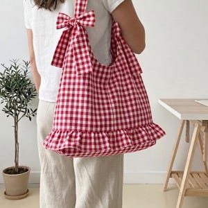 May include: A red and white gingham tote bag with a frilled hem and bow detail on the strap. The bag is carried over the shoulder. The bag is a light, casual style.