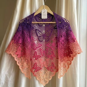 May include: A handmade crochet shawl in a butterfly design. The shawl features a gradient of colors, transitioning from purple to pink and ending in peach. The openwork design showcases butterfly motifs throughout the shawl, which is displayed on a wooden hanger.