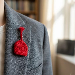 May include: A red, hand-knitted brooch in the shape of a small hat, pinned to a gray blazer. The brooch has a textured surface and a tassel at the top. The blazer has a classic design with a pocket.