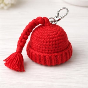 May include: A miniature red crocheted hat keyring with a silver clasp. The hat has a ribbed brim and a braided cord with a tassel. The keyring is a small version of a winter hat, perfect for attaching to keys.