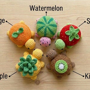 May include: A collection of five crocheted fruit-themed turtles. The turtles are designed to resemble an orange, watermelon, strawberry, pineapple, and kiwi. Each turtle has a unique color scheme and design, with the fruit names labeled above each.