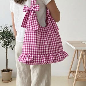May include: A large tote bag with a pink and white gingham pattern. The bag has a ruffled bottom and a bow detail on the strap. The bag is carried over the shoulder. The bag is made of a soft fabric.