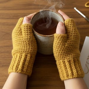 Könnte beinhalten: Senfgelbe Häkel-Fingerhandschuhe. Die Handschuhe sind am Handgelenk gerippt und bedecken die Hand bis zu den Knöcheln, wobei die Finger frei bleiben. Eine Person hält eine Tasse Tee, aus der Dampf aufsteigt.