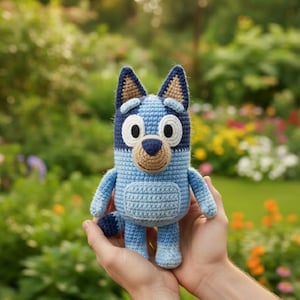 May include: A handmade crochet Bluey doll in shades of blue, tan, and white. The doll features a detailed design with a rounded body, small arms and legs, and a tail. The doll is held in a person's hands against a blurred background of greenery and flowers.
