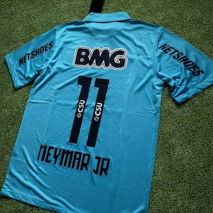 Può includere: Una maglia da calcio azzurra con il numero 11 in nero, il nome "NEYMAR JR" e la scritta "NETSHOES" stampati sulle maniche. La maglia presenta anche il logo "BMG" in bianco e nero.