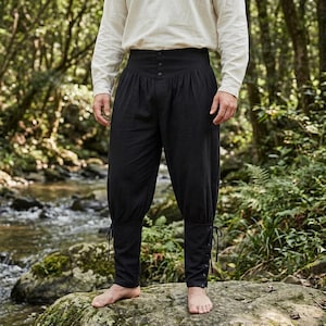 May include: Black trousers featuring a high-waisted design with three buttons at the center front, gathered fabric at the waist, and fitted lower legs with side lacing details. The trousers are paired with a long-sleeved cream-colored shirt.