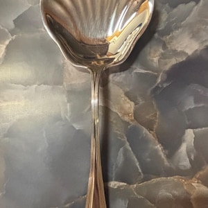 May include: A silver-toned serving spoon featuring a bowl shaped like a scalloped seashell with deep, vertical ridges. The handle is slender and elongated, transitioning into a flared base with a decorative, embossed floral motif at the tip.