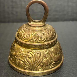 May include: A vintage brass bell with an ornate design. The bell has a looped handle and detailed relief carvings of birds, foliage, and text around the base. The bell has a tarnished, aged appearance.