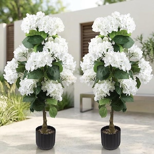 May include: Two artificial hydrangea trees in black pots. Each tree features a brown trunk, green leaves, and large clusters of white flowers. The pots have a ribbed design. The trees are placed on a light-colored patio.