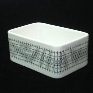 May include: A rectangular white ceramic dish with a black geometric pattern around the sides. The pattern consists of vertical lines with leaf-like shapes at the top, connected by a series of diagonal lines.