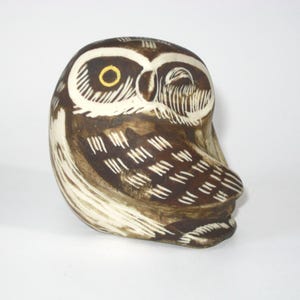 May include: A ceramic owl figurine with brown and white painted details. The owl has a yellow eye and is looking to the right.